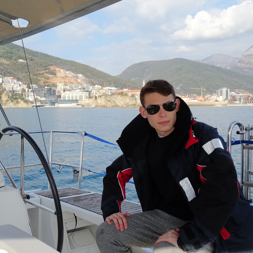 Zdjęcie profilowe Rafał Szostecki, poważny, siedzący przy sterze jachtu, oparty o kolana, w tle miasto na lądzie